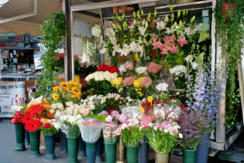 A vibrant outdoor flower display featuring a variety of fresh floral arrangements and bouquets. Prominent among them are white lilies with large, open petals and long, green stems, arranged at the top