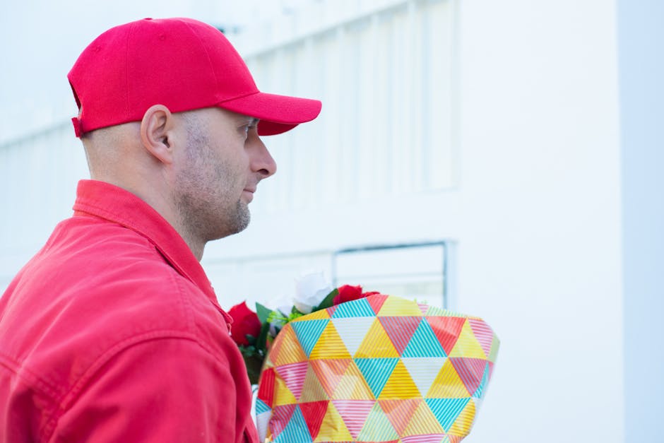 A delivery person dressed in a red jacket and matching cap is holding a vibrant floral gift wrapped in yellow, blue, red, and green geometric patterned paper, with a red ribbon. The person's profile s