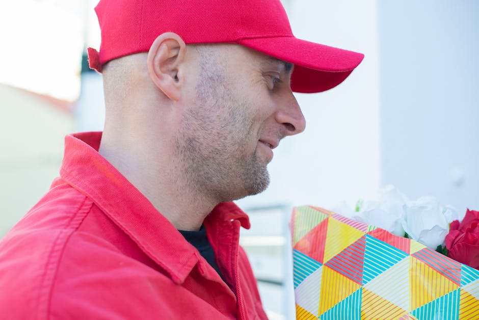 A man wearing a red cap and matching red jacket holds a floral gift wrapped in colorful geometric-patterned paper. The arrangement features fresh white and pink flowers, including roses, with visible 