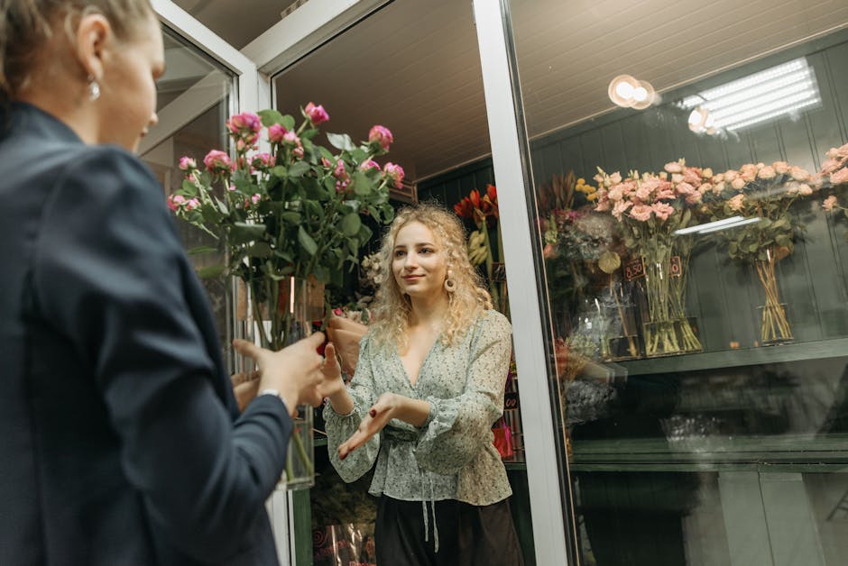 A female florist with curly blonde hair, wearing a patterned blouse, is handing a bouquet of pink roses wrapped in brown paper to a customer inside a flower shop. The bouquet features fresh pink roses